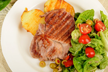 two pieces of grilled potato and grilled meat next to lettuce with tomatoes and olives on a white plate