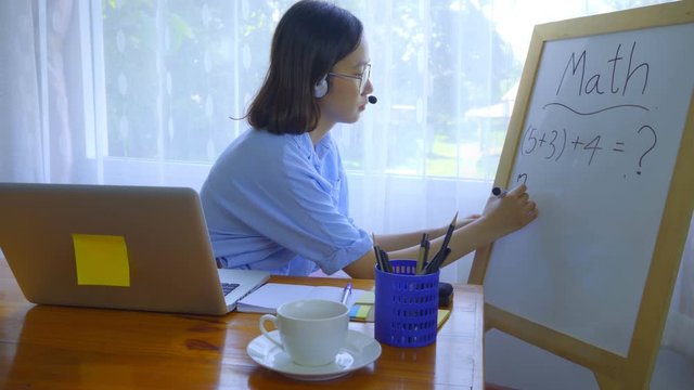 An Asian woman is teaching online via a laptop via webcam.