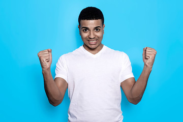 Fototapeta premium Overjoyed black man raises clenched fists, feels energized while gesturing happily against blue wall