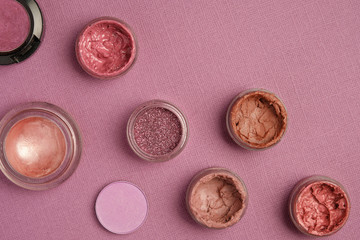 Eye shadow, glitter and lipstick in nude and pink shades are placed diagonally on a dirty pink textured background