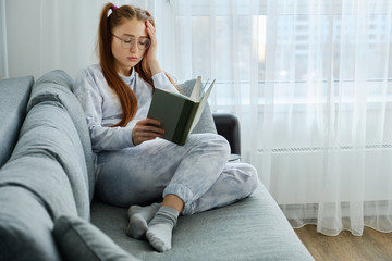 A red-haired girl with tails in pajamas lies on the sofa, legs crossed and reading a book