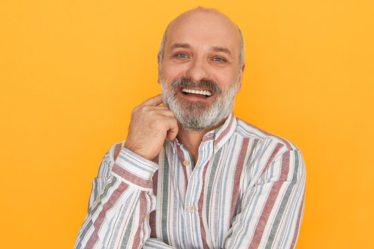 Studio image of easygoing sociable attractive senior man with bald head and thick beard holding hand at his face, looking at camera with broad smile, telling jokes, having fun. Joy and happiness