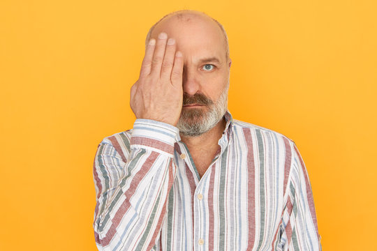 Studio image of serious senior man with baldness and gray thick beard covering one part of face with palm, having his eyes been checked during vision test, suffering from long sightedness