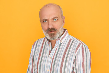 Obraz premium Horizontal shot of grumpy displeased senior man with thick gray beard and bald head staring at camera having suspicious look, frowning, not trusting you. Human facial expressions and reaction