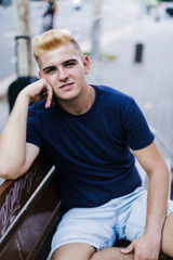 Portrait of a young teenage male with blonde bangs sitting on a bench in the street while looking at the camera