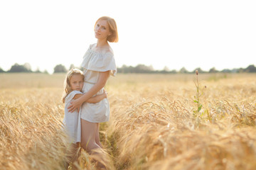 Naklejka na ściany i meble A beautiful red-haired young mother and her daughter hug each other in a wheat field on a Sunny day.