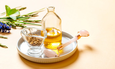 Cosmetic oil, herbs and Rose quartz facial roller on a beige background. Facial skin care at home, anti-aging and lifting therapy. Pink gua sha facial massager tools and oil. Natural home skin care 