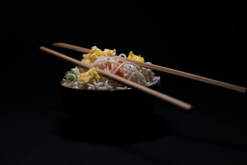 A bowl of asteamed noodles and vegetables decorated with veggies and chopsticks in a black copy space background. Food photography.