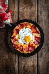 Sourdough breakfast pan pizza with pepperoni and fried egg in a skillet on wooden surface