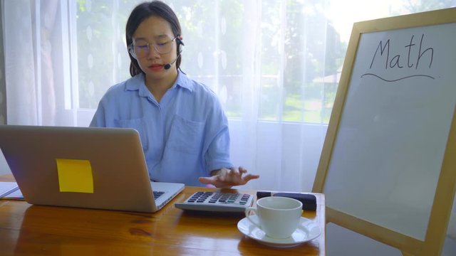 An Asian woman is teaching online via a laptop via webcam.