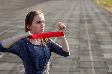 Young slim woman in sportswear doing squats exercise with rubber band on a black coated stadium track