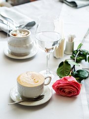 Restaurant. A white tablecloth, a Cup of freshly brewed cappuccino and a glass of water. A pink rose is lying next to it. In the background, cane sugar. Breakfast, lunch or dinner. Menu.