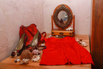 Composition of shoes, a red dress, shells, a mirror, jewelry, and dried flowers on a wooden table.