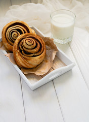 Two yeast rolls cinnamon rolls lie on a wooden tray and a glass of milk on a white wooden background.