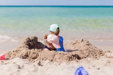 Two sister girls in swimsuits and caps playing on the beach with toys and sand on a sunny summer day. Fun family vacation at the sea. Activity outdoors
