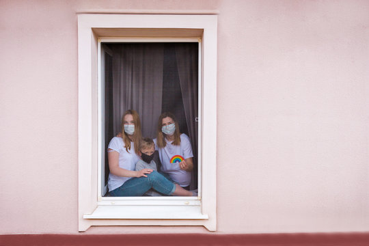 Rainbow In The Window. Thank You NHS, Promotion In Support Of Doctors In The Coronavirus Pandemic, Covid 19. Family In The Window Of The House In Masks And A Rainbow In  Hands. National Health System