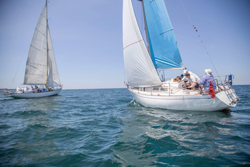Fototapeta premium Two white motor yachts with raised sails take part in the regatta. A strong wind tipped the ship. Waves, a small wave amplify emotions at competitions. Boat crews are busy navigating and chasing. Boat