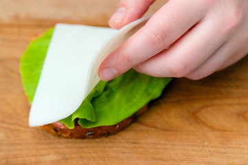 Close up of hand placing cheese on bread slice with salad leaf on wooden cutting board. Making sandwich
