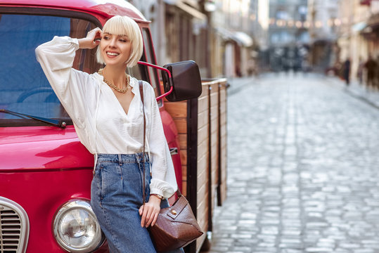 Street style photo of happy smiling fashionable woman wearing trendy white blouse, high waist jeans, with brown faux croco leather textured shoulder bag. Model posing in street of European city