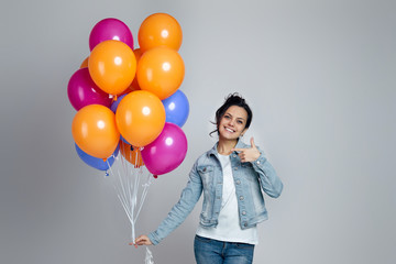 happy young caucasian woman in denim pointing at bright colorful air balloons isolated on gray background. birthday party.