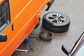 Removed wheel and brake drum are lying on the road next to the orange minibus on a hydraulic Jack, overheating the jammed car brakes on the way