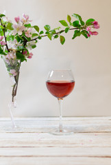 Glass of pink wine and apple tree branches with pink flowers in a vase on white wooden table