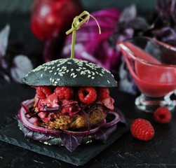 Fashion black burger with raspberries and  berry sauce. On a dark background. Inside the cutlet, red onions, purple basil, red cabbage and raspberry homemade vinaigrette. 