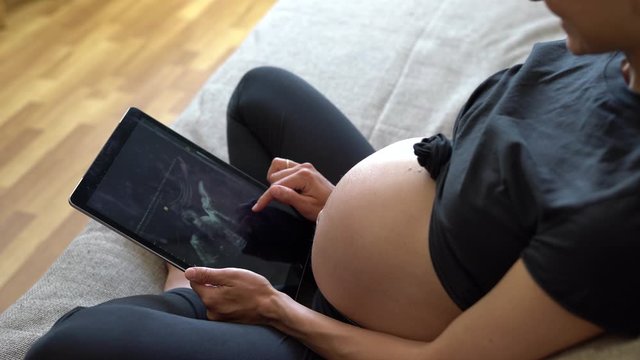 Video of beautiful young pregnant woman looking ultrasound of her baby on digital tablet while lying on bed
