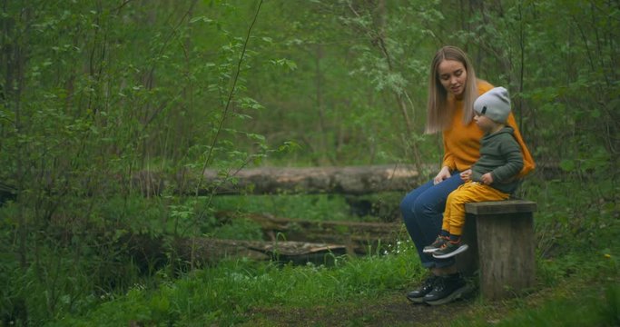 Mother and son sitting on a log of a tree in the forest near the stream talking and pointing ahead. Look ahead. To consider and explore. Mother tells her son and introduces him to nature.