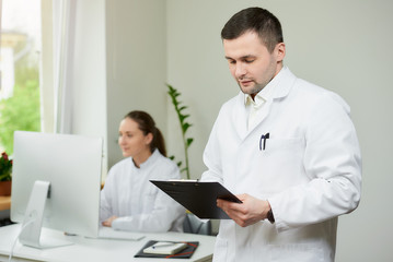 Fototapeta premium A surgeon with bristles in a white coat holds a black clipboard with documents and a pen in a hospital. A female doctor sitting in front of the desktop computer in the corner of the room. 