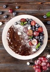 chocolate cake with fresh berries, red grapes and blueberries, mint, coconut crumbs, powdered sugar and marshmallows on a white plate on a wooden