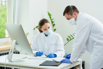 A surgeon and a radiologist in medical face masks make a diagnosis of a patient in a hospital. A female doctor sitting in front of the desktop computer, a therapist stays near her.