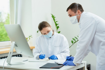 Obraz premium A surgeon and a radiologist in medical face masks discuss a diagnosis of a patient in a hospital. A female doctor sitting in front of the desktop computer, a therapist stays near her.
