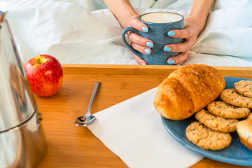 Breakfast in bed with fruits and pastries on a tray and womans hands hold cup of coffee