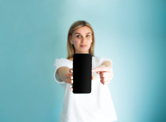 Naklejka premium Blank screen smartphone shown by young woman on a blue background. Copy space mockup, pointing on a screen