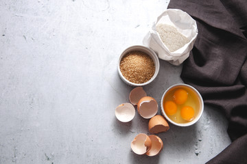 Obraz na płótnie Canvas Dough baking ingredients: a bag of flour, eggs, egg with yolk in a bowl, brown sugar with rolling pin on light grey concrete background. Top view, flatlay, copy space. Recipe Mockup