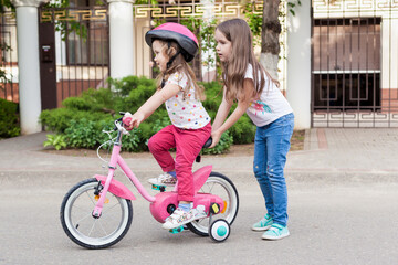 Children learning to drive a bicycle on a driveway outside. A little girl teaches her sister to ride on asphalt road near the house . Active healthy outdoor sports for young kids. 