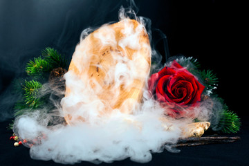 Wooden stump (cut log), red rose flower, different peppercorns, fir branches, bourbon vanilla pods and ginger root with smoke on a black background. Selective focus.