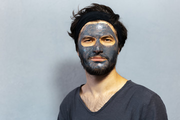 Portrait of young disheveled guy with beauty facial mask on background of grey background.