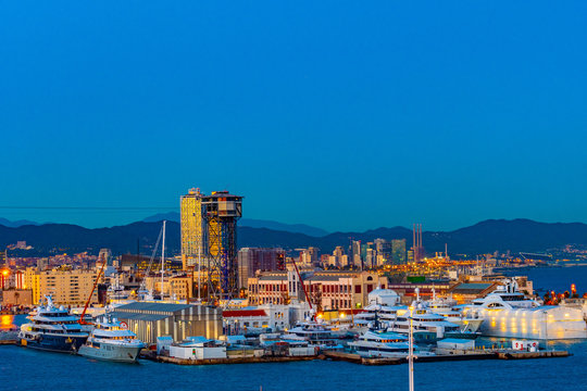 Barcelona, Spain. Night city skyline in Catalonia. Modern architecture and yachts docked on coast near Torre Jaume I aerial lift pylon steel truss tower of Port Vell Aerial Tramway.