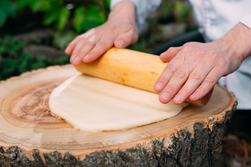 A restaurant chef in uniform rolls out dough on a stump in the woods.