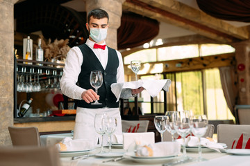 A waiter in a medical protective mask serves the table in the restaurant.