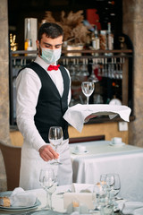 A waiter in a medical protective mask serves the table in the restaurant.