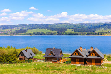 Fototapeta na wymiar Traditional houses on green meadow and view of Czorsztynskie lake near Falsztyn village on sunny spring day, Pieniny Mountains, Poland