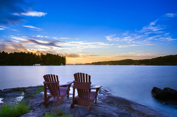 Fototapeta na wymiar Long exposure of two Muskoka chairs sitting on a rock formation facing a calm lake at dusk in cottage country.