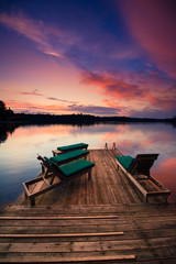 Lounge chairs sitting on a wooden dock facing a calm lake at sunset. 