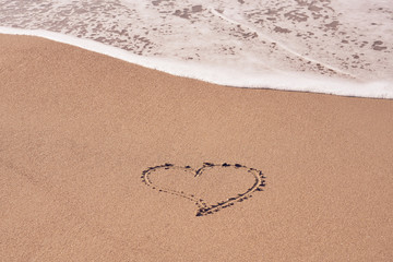 A heart symbol written on a sandy beach with foam and water background. Valentines day concept.
