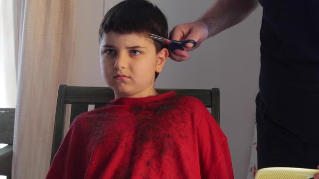 Hair cutting at home. Epidemic of coronavirus infection COVID-19. Hairdressers are closed.A serious child on a haircut by dad. A boy in red clothes is scissored by cutting bangs on his head.