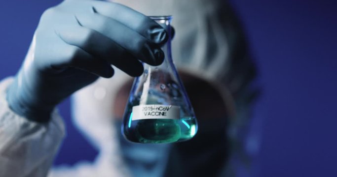 Close up of glass flask with liquid vaccine against coronavirus in hands of male Caucasian laborant in full protected costume, mask, goggles and gloves. Laboratory testing of vaccines.