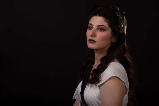 Close-up portrait of a archer woman in white dress on black background. Greek goddess Artemis. Studio photo shoot. Professional makeup and hairstyle for a brunette with long dark hair. Young woman